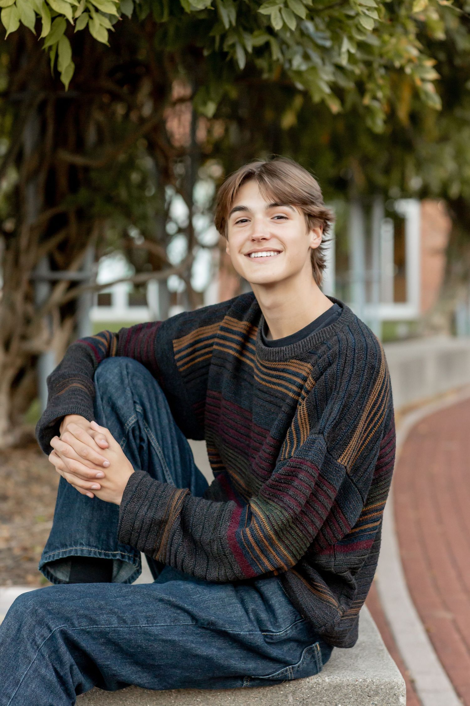 Smiling teenage boy in striped sweater sitting on a ledge outdoors near brick building.