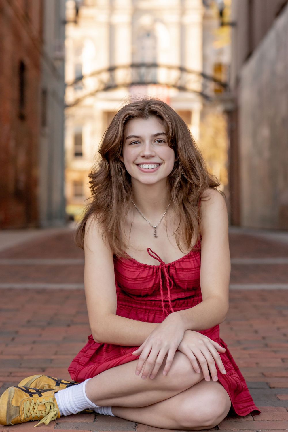 Smiling teenage girl in red dress sitting cross-legged on brick pathway in urban setting.