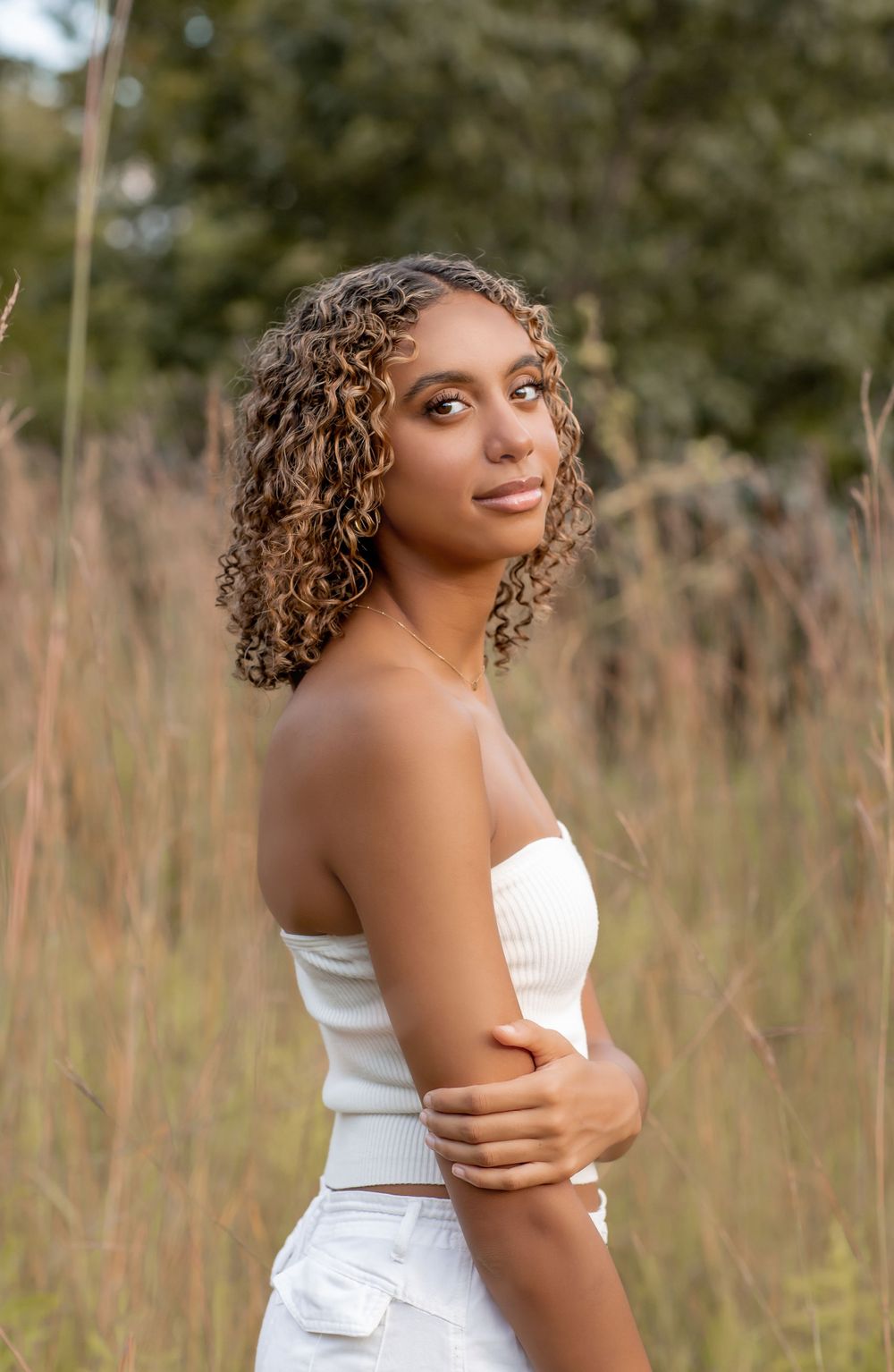 Young woman with curly highlighted hair in white tube top posing in tall grass field.
