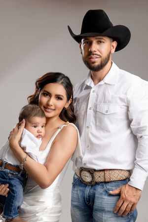 Smiling woman in a white satin dress holding a baby beside a bearded man in a black cowboy hat, white button-up shirt, and decorative belt.