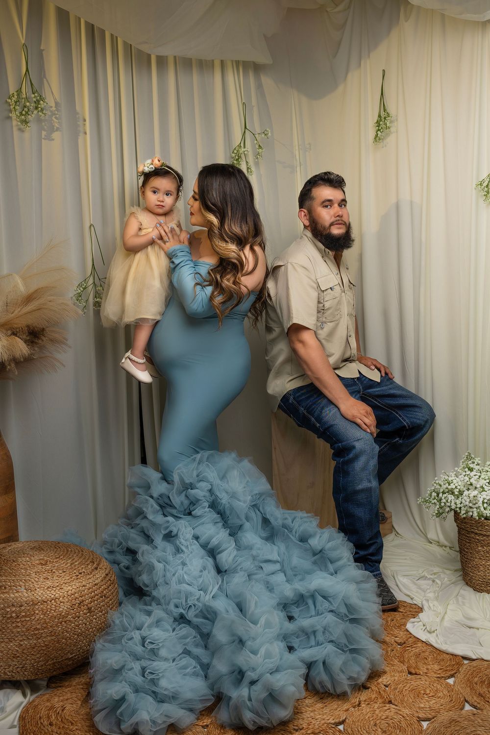 A family poses in front of a cream curtain backdrop with a woman in a blue ruffled gown holding a small child.