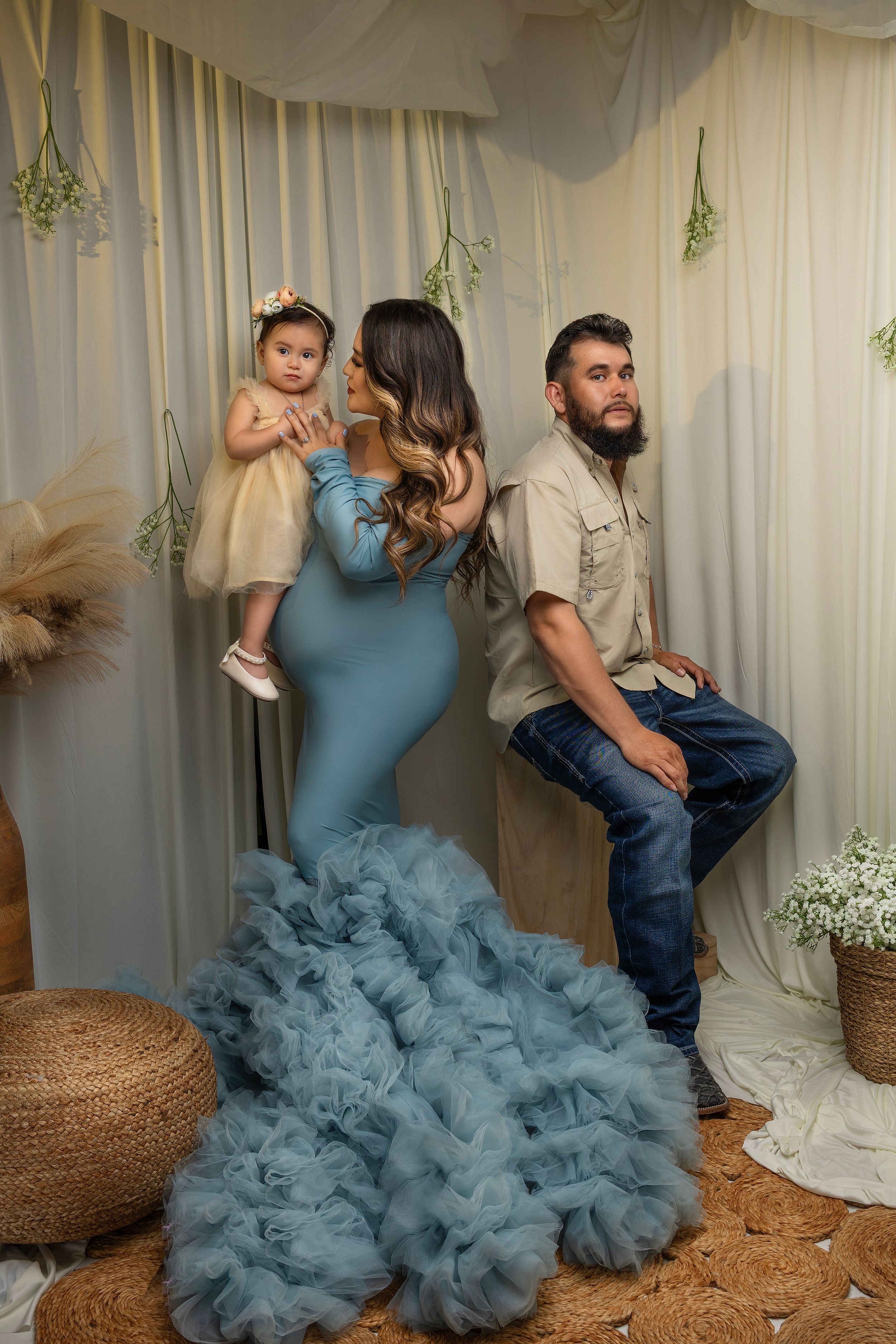 A family poses in front of a cream curtain backdrop with a woman in a blue ruffled gown holding a small child.