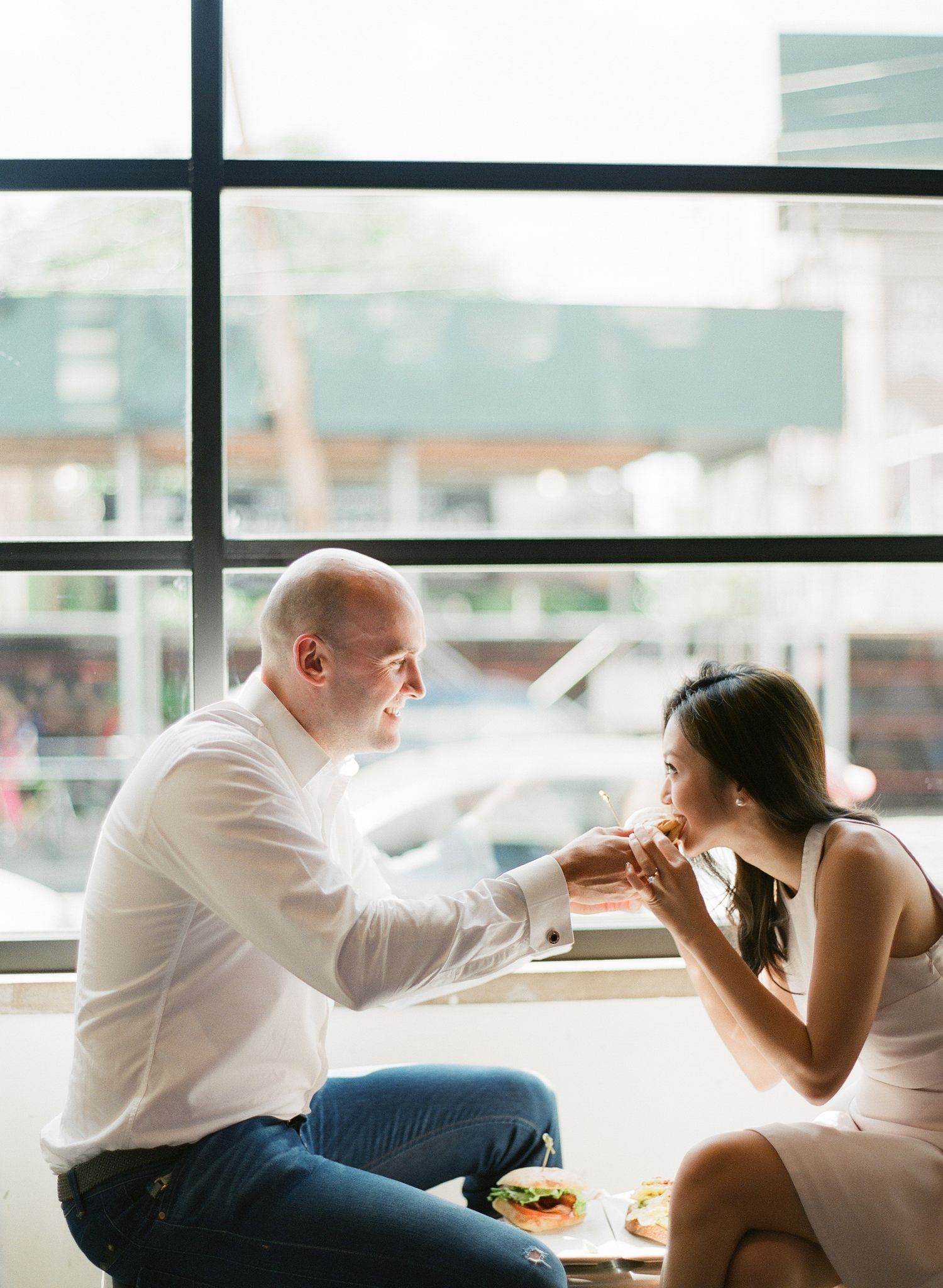 Couple sharing a romantic moment over food by a large window in a bright, modern restaurant setting.