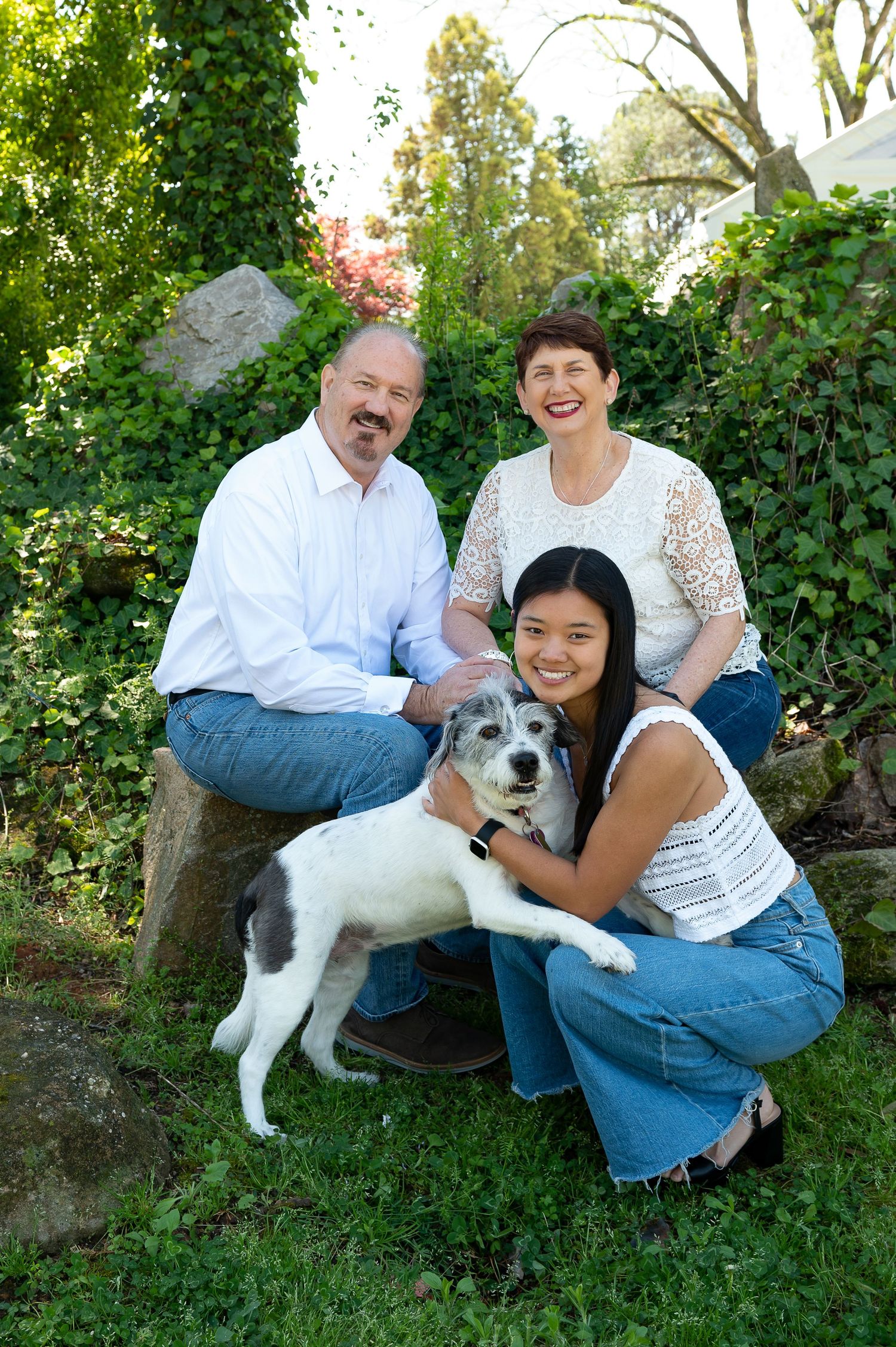 Family outdoor portrait with black and white dog in green garden setting.