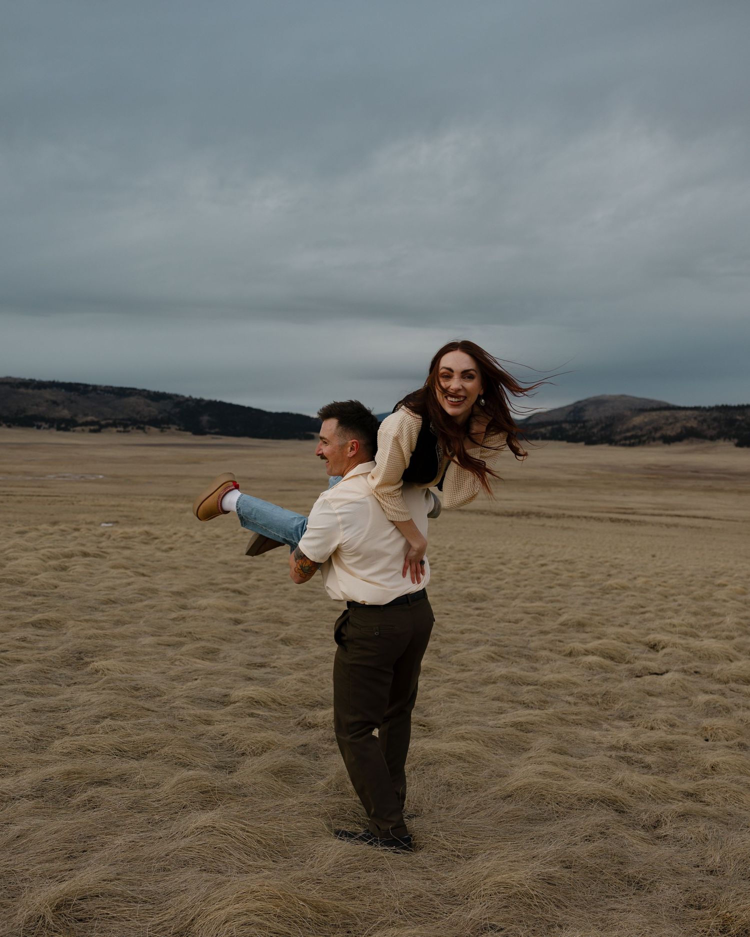 A couple walks hand in hand along a scenic beach with mountains in the distance at sunset.