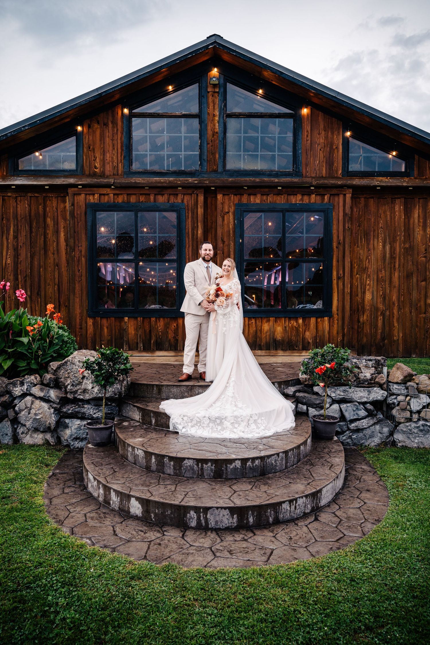 Newlyweds pose on circular stone steps in front of rustic wooden barn venue with string lights.