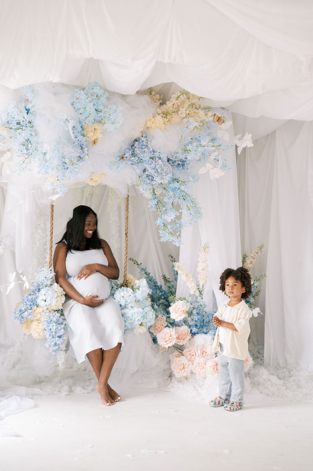 Dreamy maternity photo session featuring soft white fabric draping, blue and peach floral decorations in an ethereal setting.