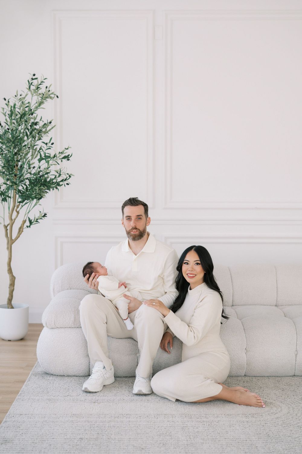 Couple sits together on white couch in bright minimalist room with potted tree in corner.