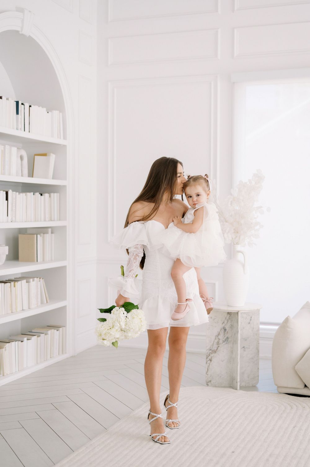 A white minimalist room featuring bookshelves and decorative pampas grass, with soft natural lighting streaming through windows.