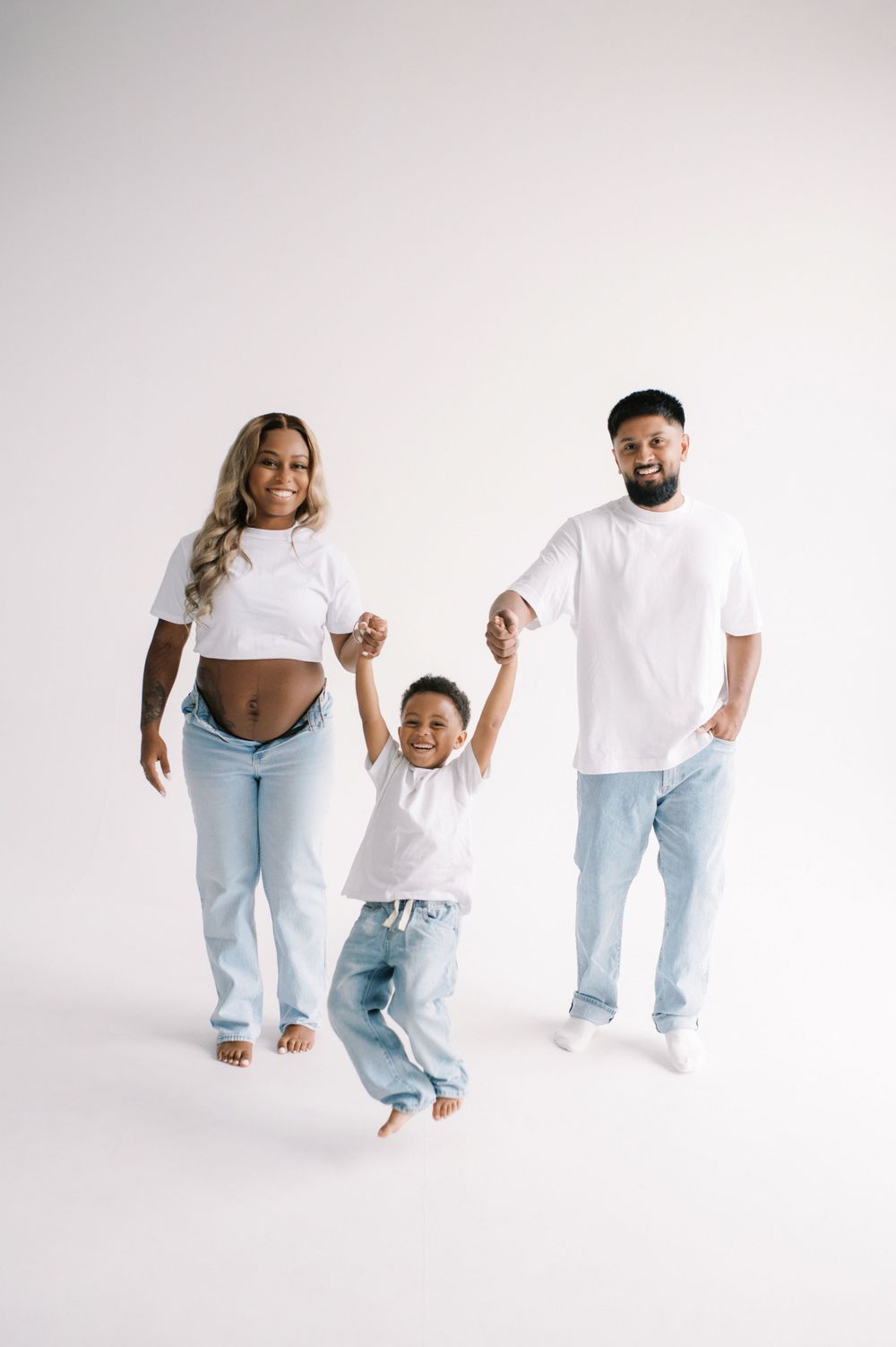 A happy family wearing white tops and light denim jeans jumps playfully against a white backdrop in a studio setting.