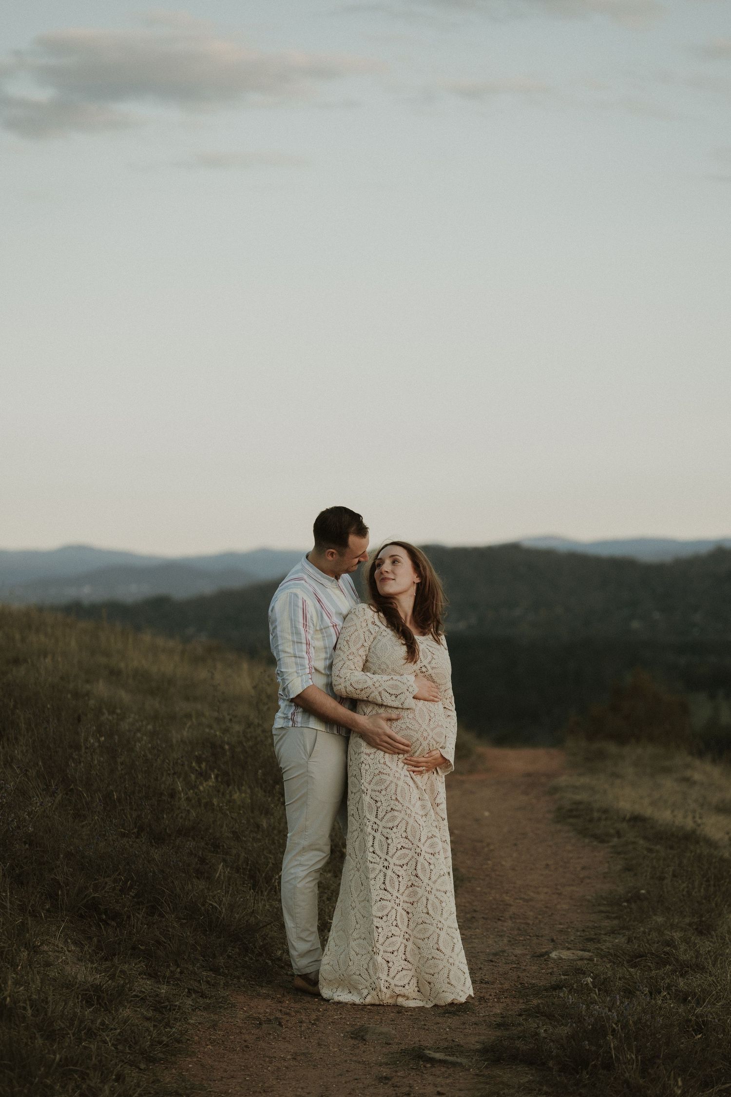 Pregnant woman in vintage lace dress stands romantically with her partner, hilltop at sunset with Canberra mountains in the background.