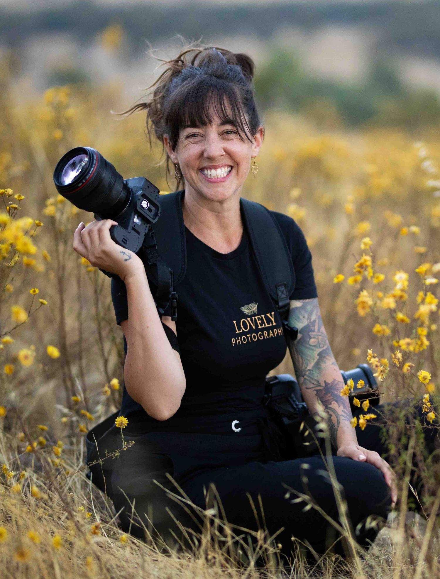 A photographer in a pink tank top and light jeans holds a professional camera while standing against a textured wall.