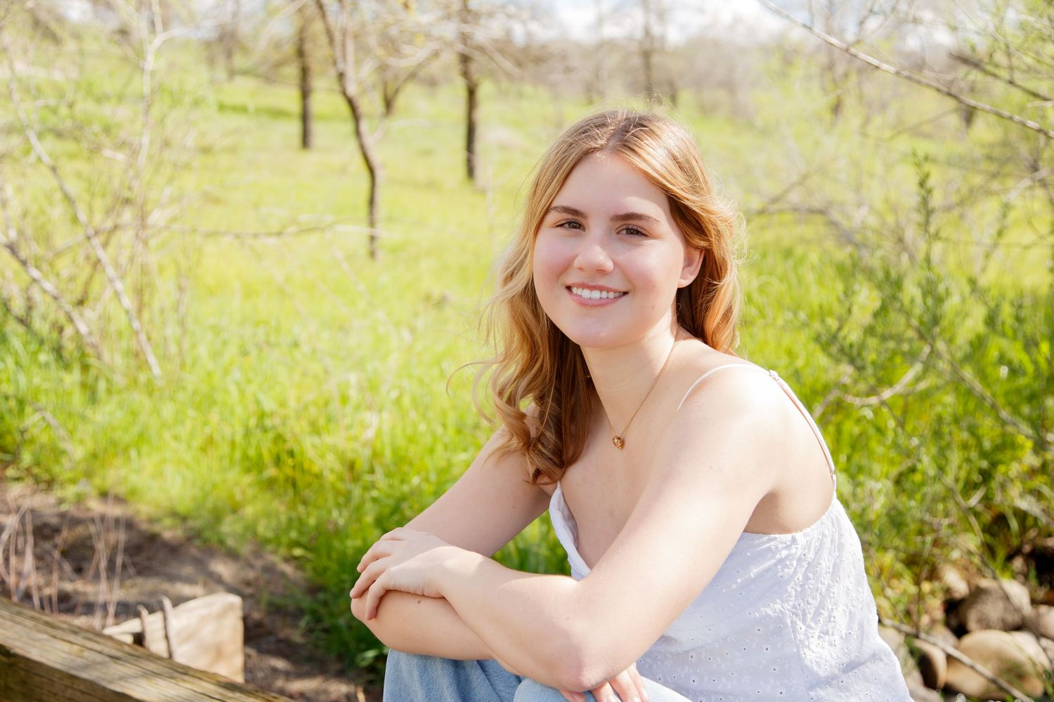 Young woman smiling outdoors, leaning on wooden fence with lush green trees in sunny spring setting.