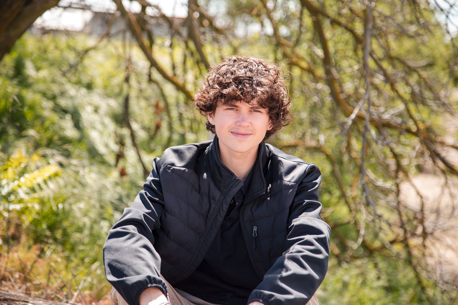 A teenage boy wearing a black jacket sits outdoors among trees and foliage in natural lighting.