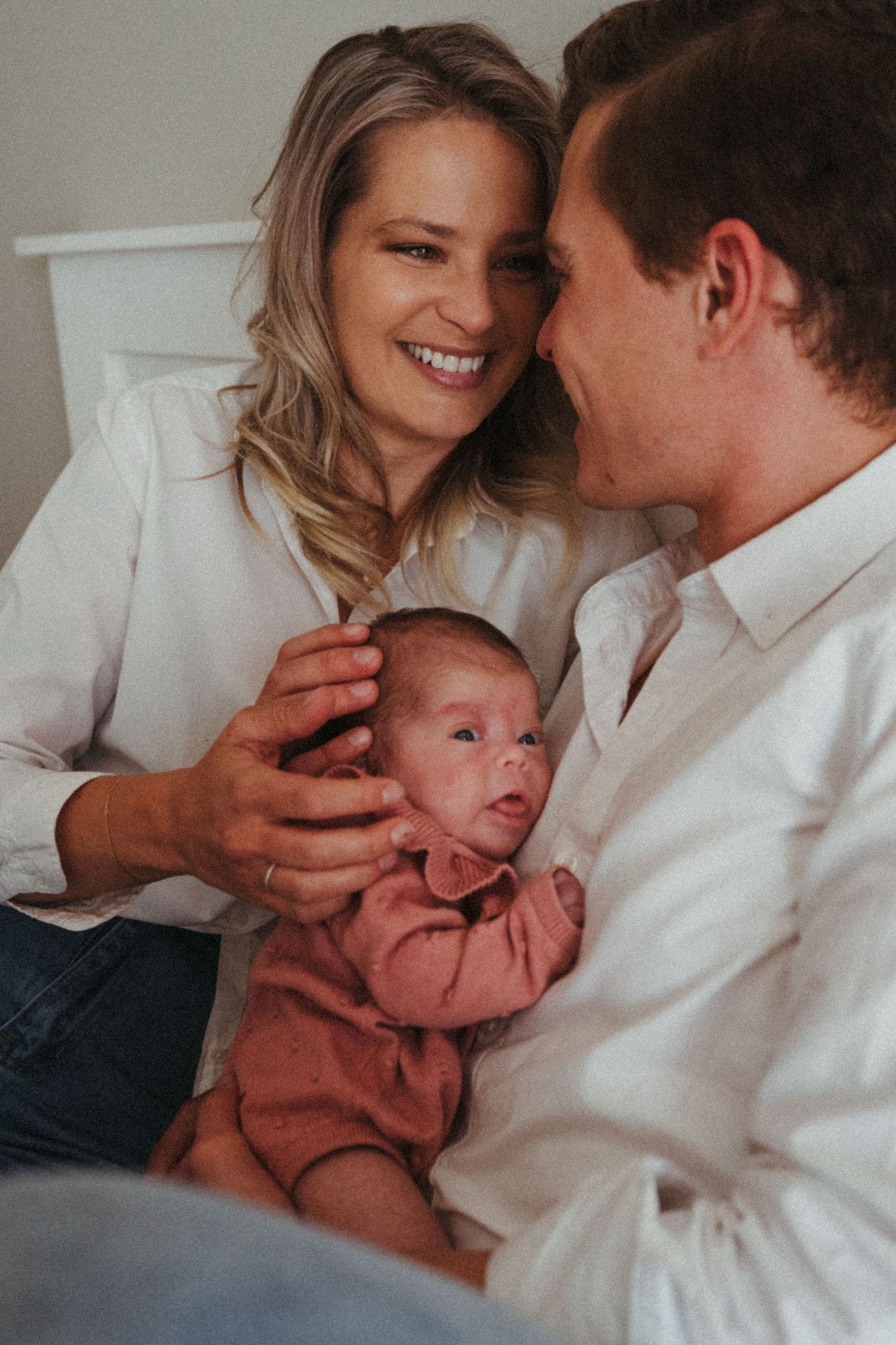 Parents sharing loving moments with their newborn baby on white bedding.