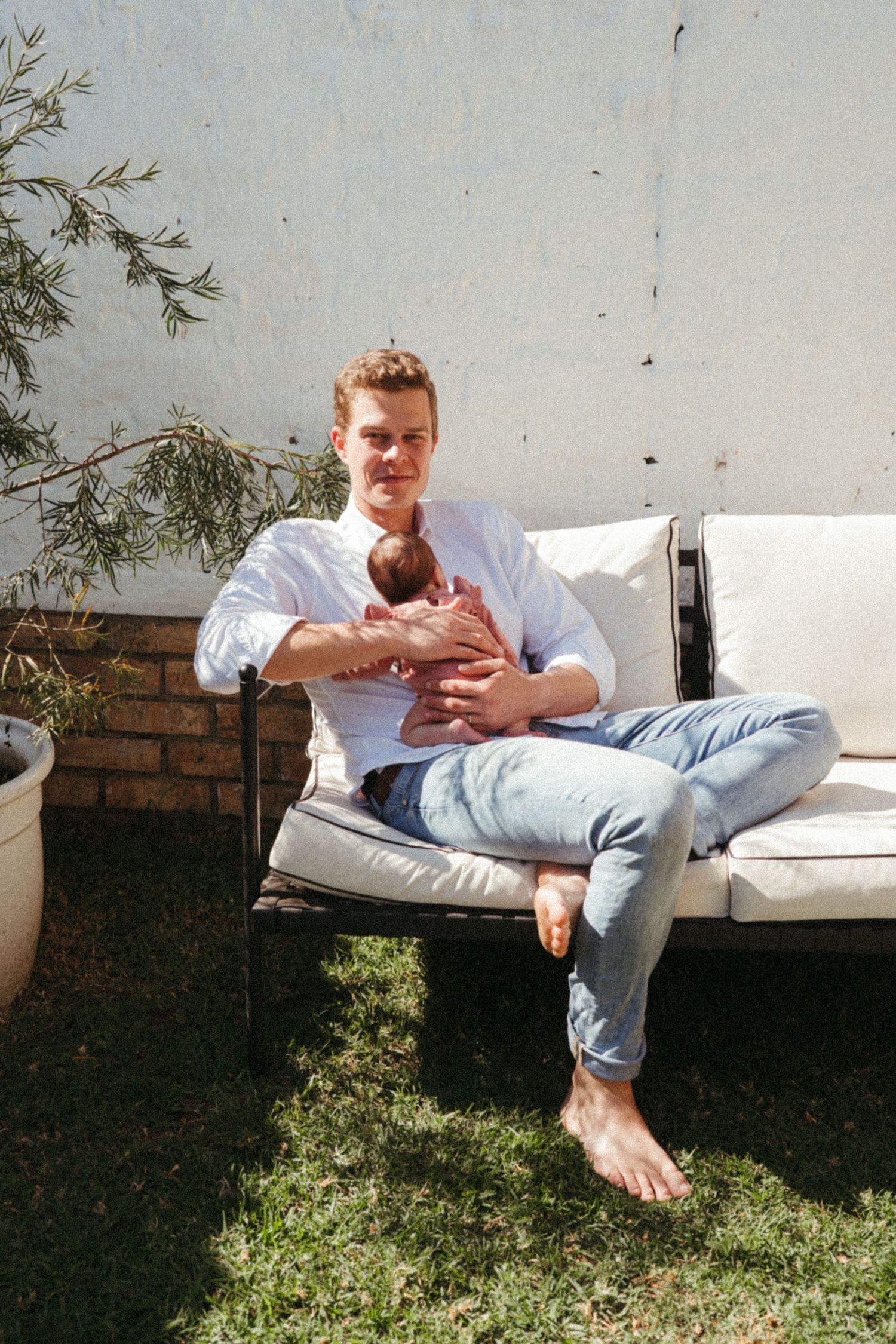 Person lounging casually on white outdoor furniture against a brick wall with greenery.
