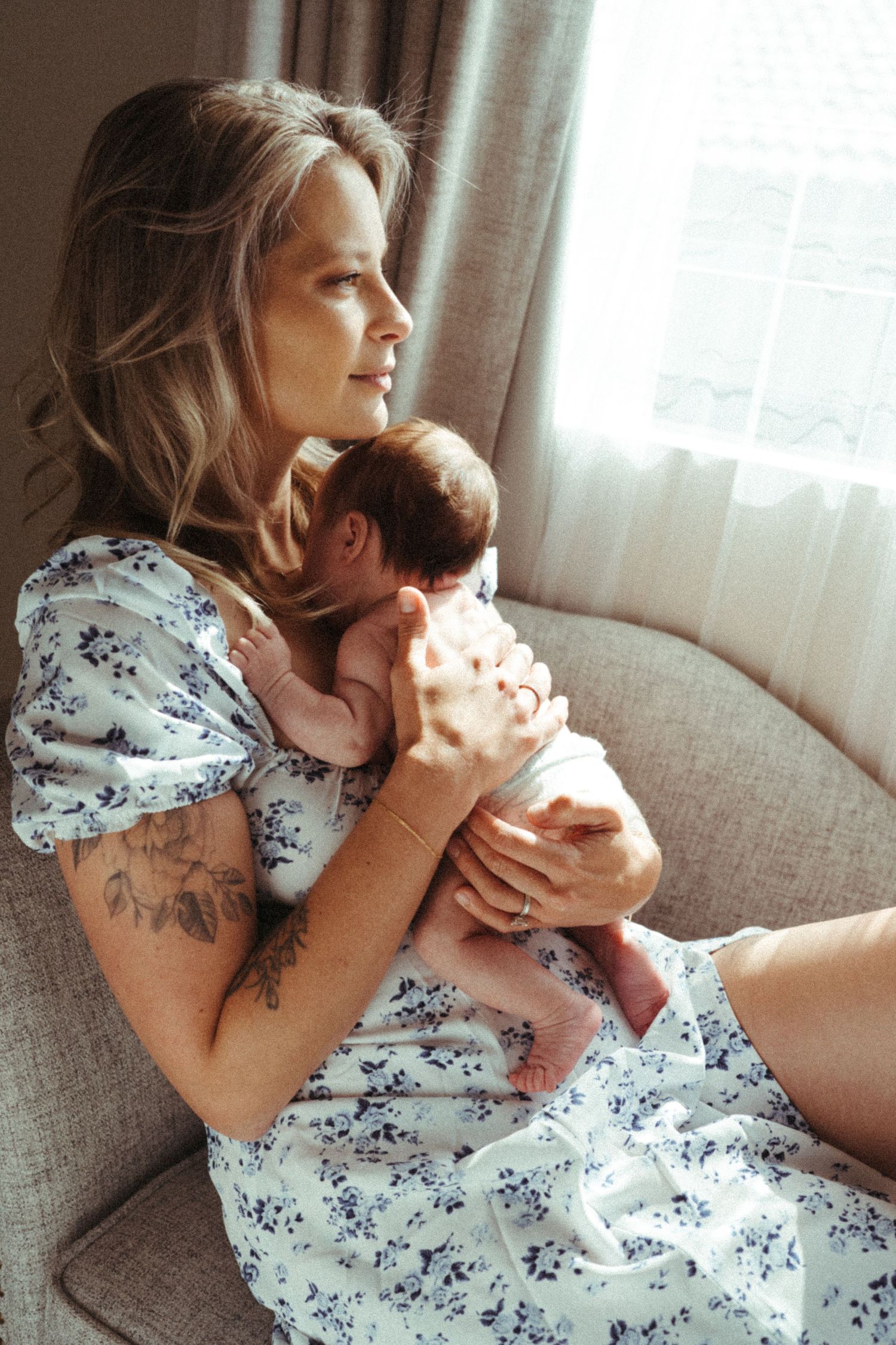 Person in floral dress relaxing with pet on grey couch near window.