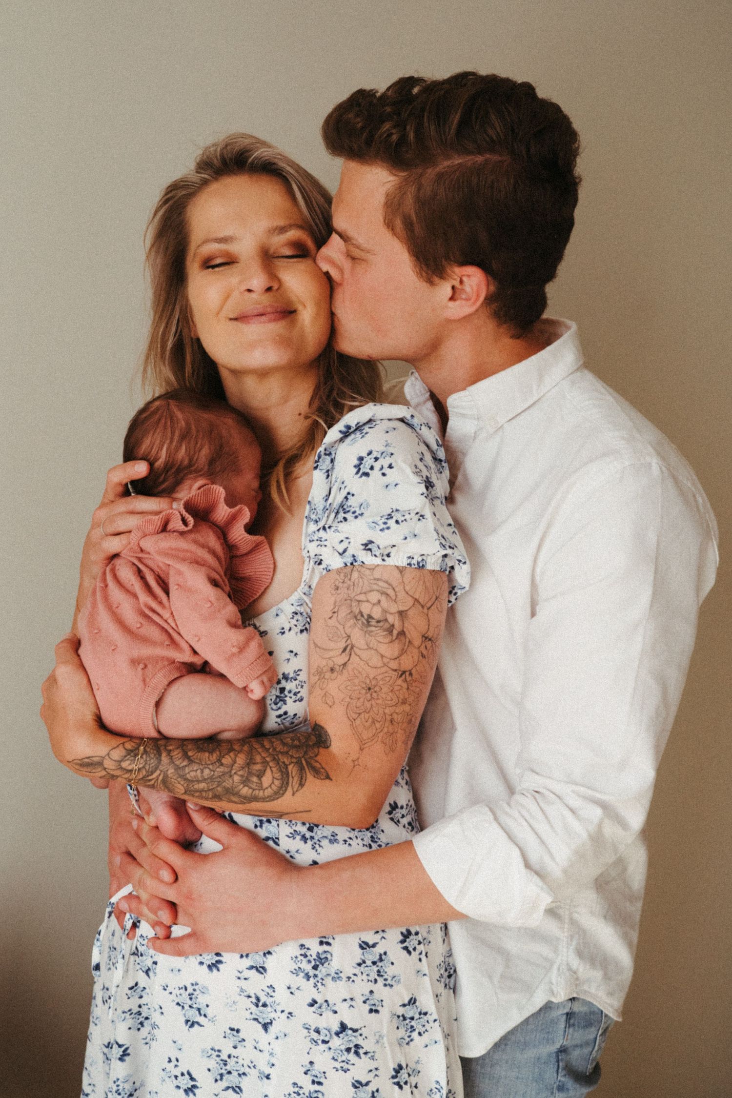 Parents in white and floral dress share tender moment while holding newborn in pink outfit.