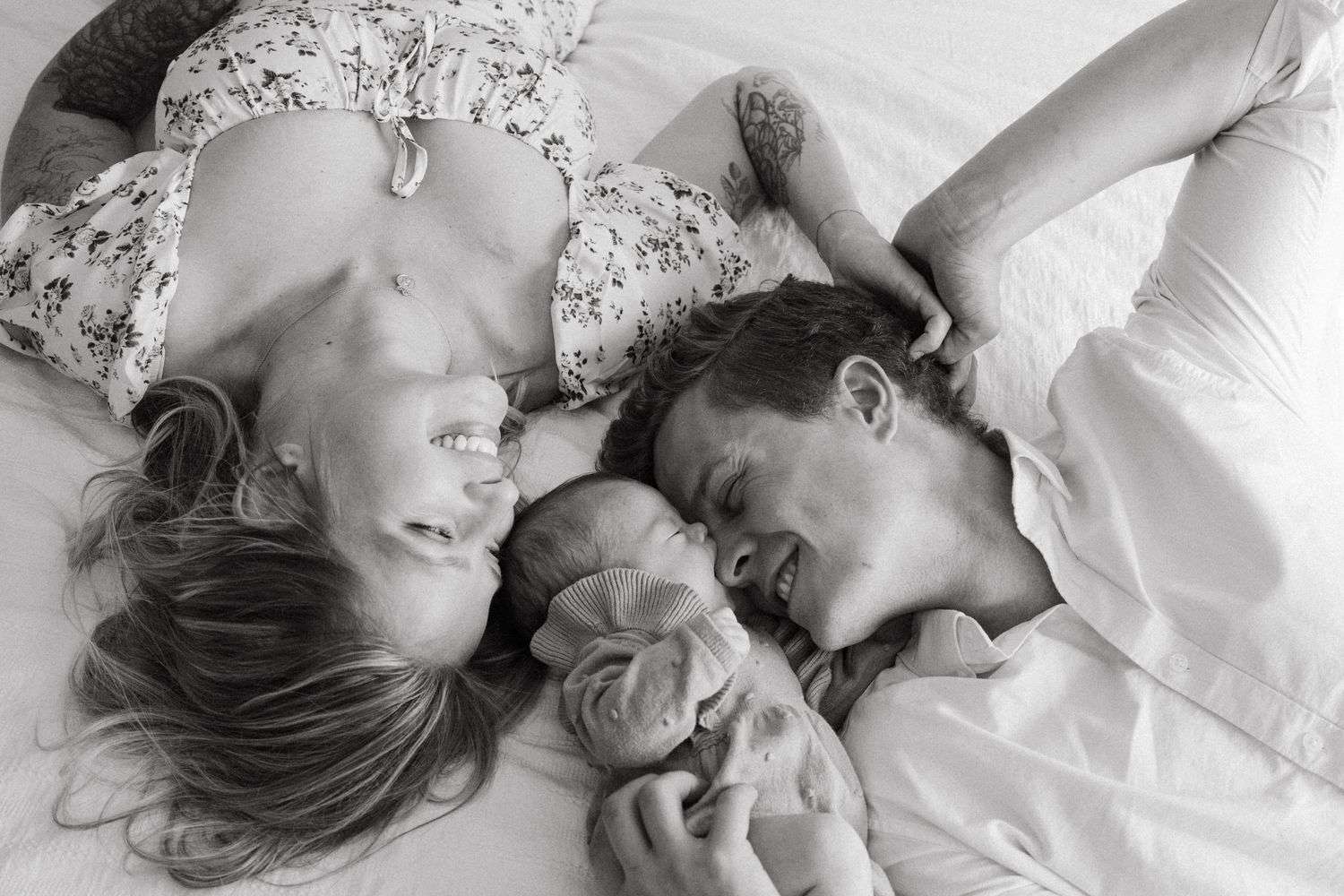 Black and white photo of parents lying with newborn baby between them on white bedding.