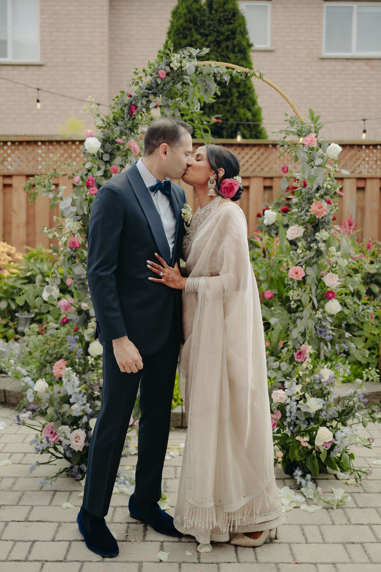 A couple shares a romantic moment under a floral arch with pink and white roses in a backyard garden setting.