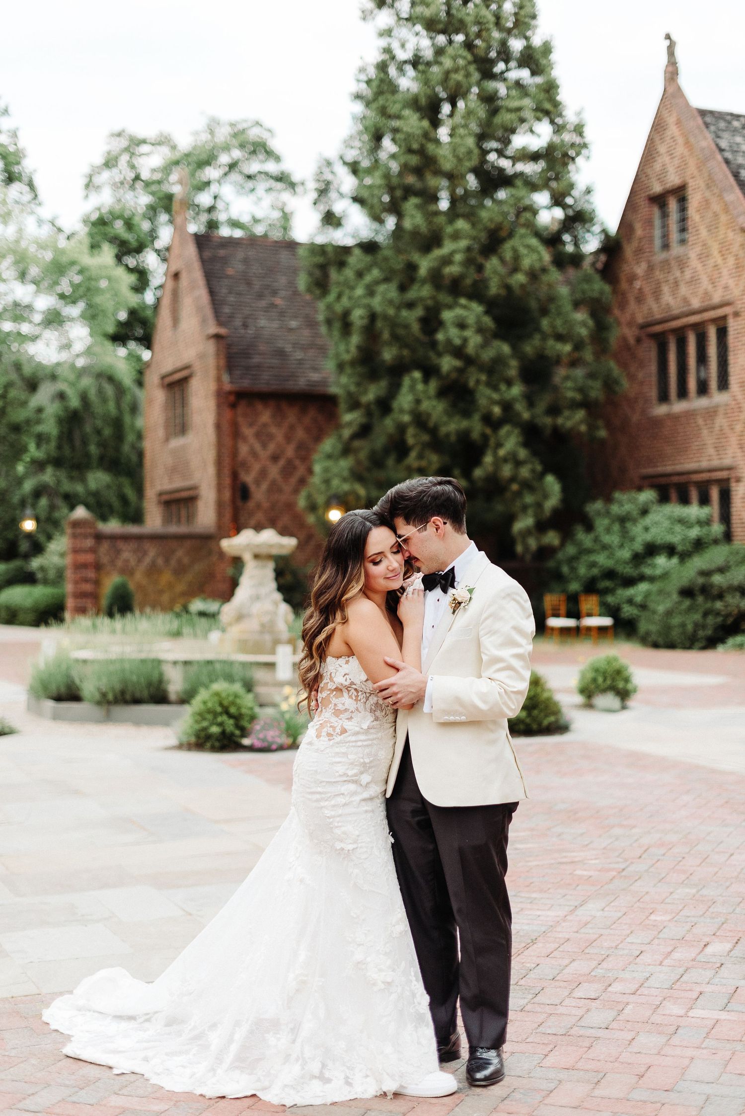 Romantic couple in wedding attire share a tender dance moment on brick courtyard with gothic chapel buildings in background.