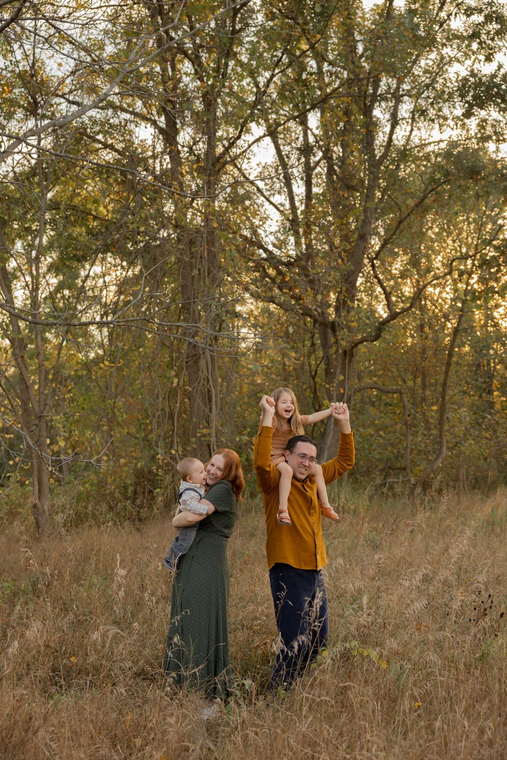 a family in fall attire dance joyfully together in a golden autumn field surrounded by trees at sunset at cranberry lake park in Rochester