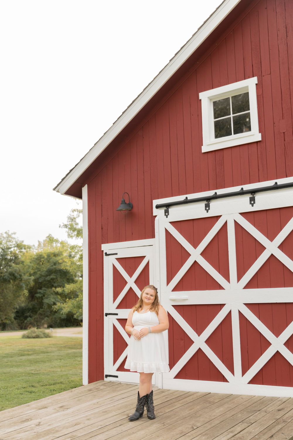 teenage girl in a white dress and cowboy boot smiling for a photo in front of a red barn in Oakland township