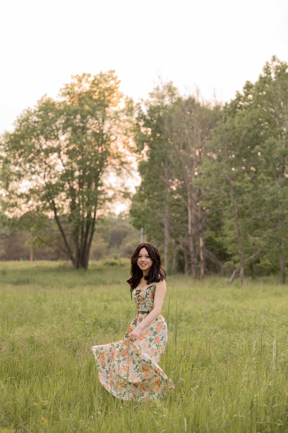 high school senior twirls in a meadow with tall grass and trees in the background during golden hour at cranberry lake park in Rochester MI