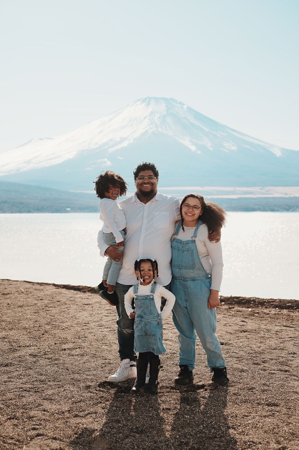 A happy family wearing matching denim overalls poses together on a beach with a snow-capped mountain in the background.