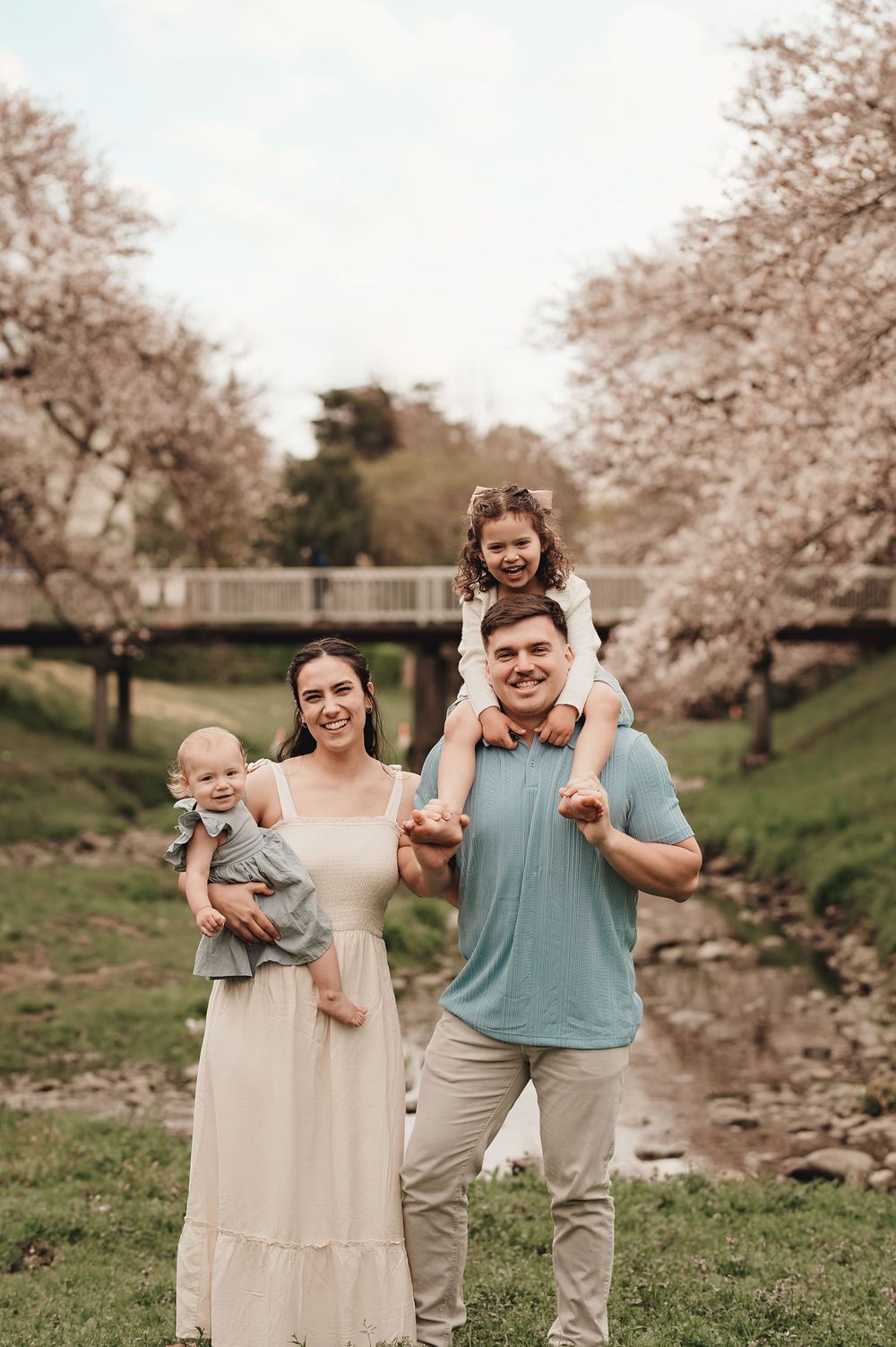 A family stands together on green grass with blooming cherry trees and a wooden bridge in the background during spring.