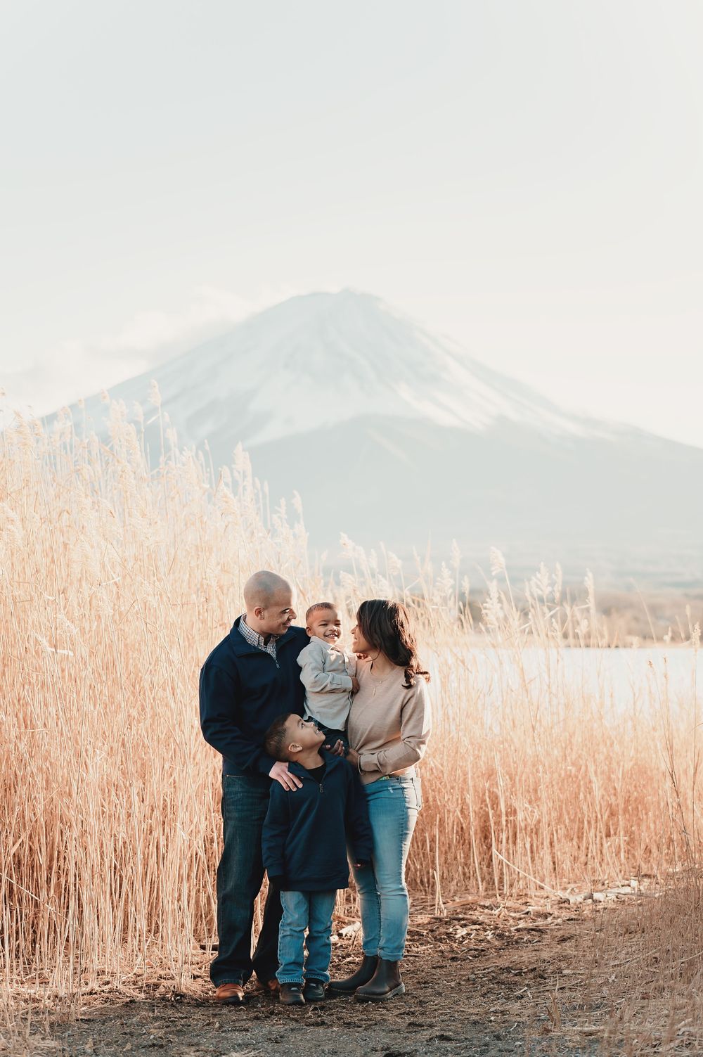 A family shares tender moments together in front of tall wheat fields with mountains in the background at sunset.