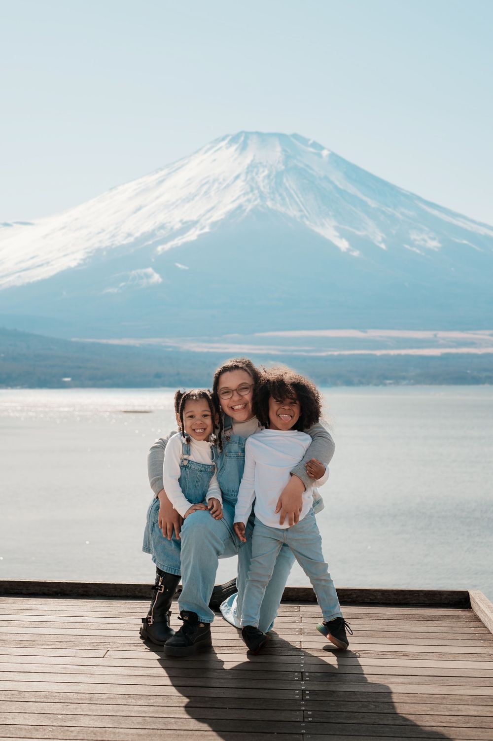 Three people pose together on a wooden deck overlooking Mount Fuji and a serene lake in Japan.