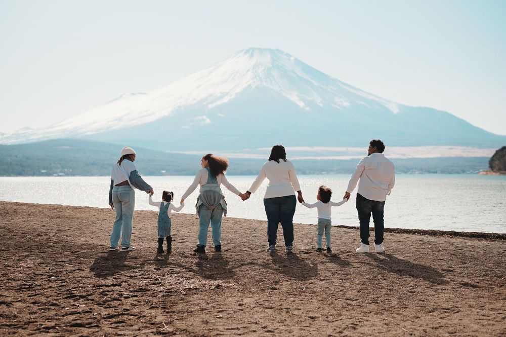 A group stands holding hands on the shore of Lake Yamanaka with Mount Fuji rising majestically in the background.