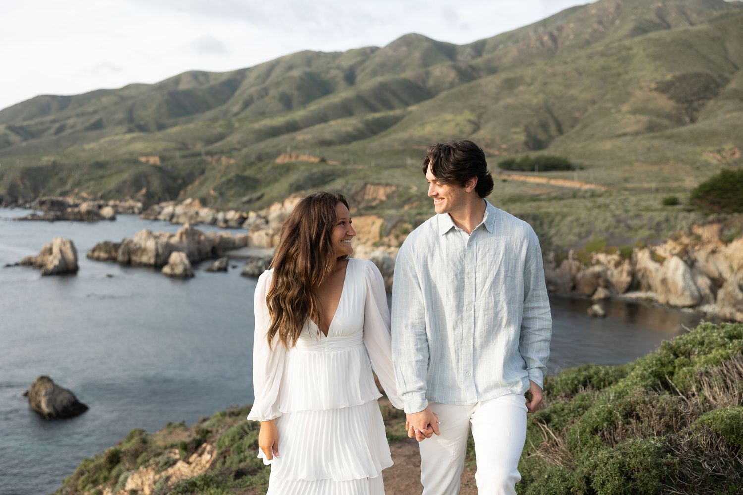 A couple shares a romantic kiss on a sandy beach, the bride in a white gown and groom in a blue suit.