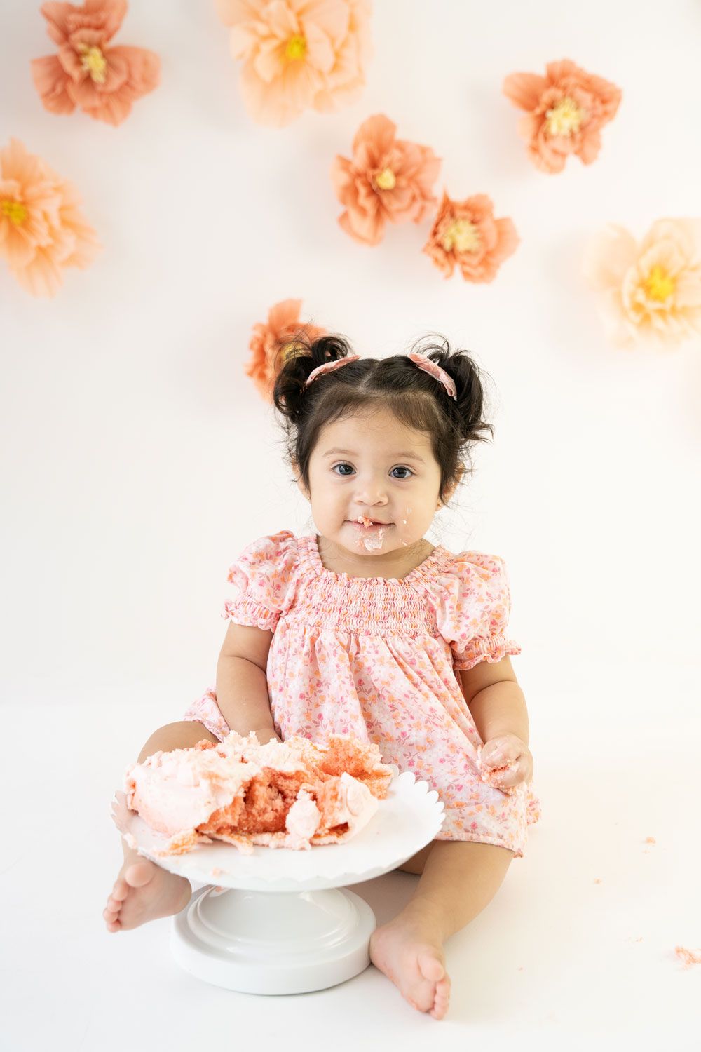 Sweet cake smash photography setup with peach floral backdrop and toddler in pink floral dress sitting with dessert.