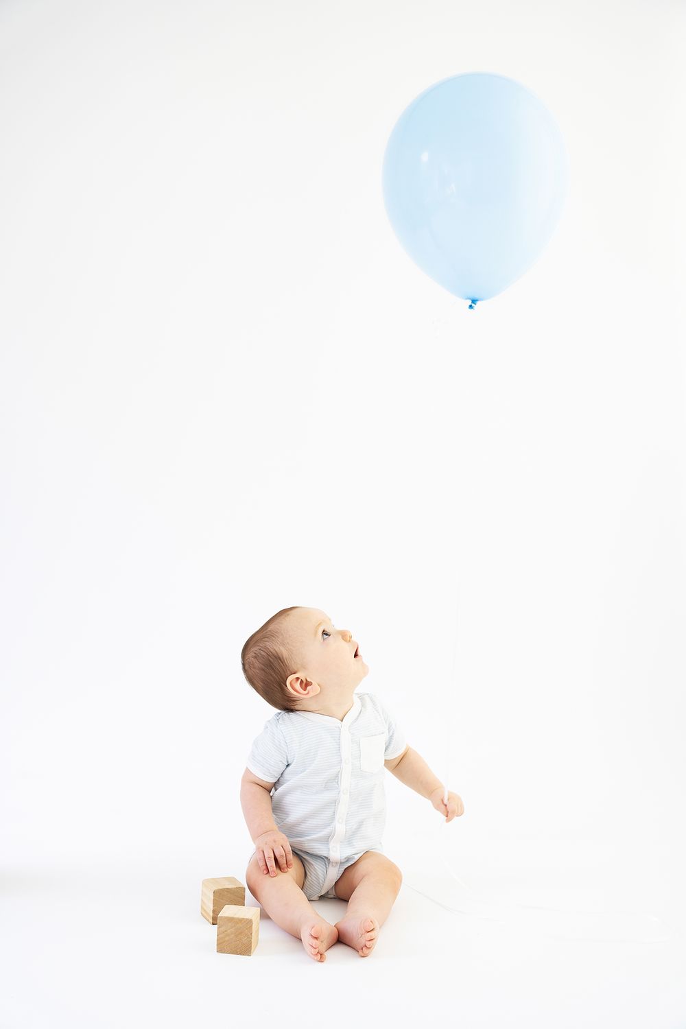 Baby in white onesie sits on floor looking up at floating blue balloon against white background.