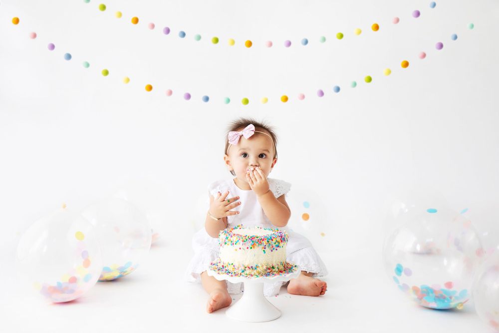 A small child in white sits on the floor eating cake with colorful party decorations and bunting hanging overhead.