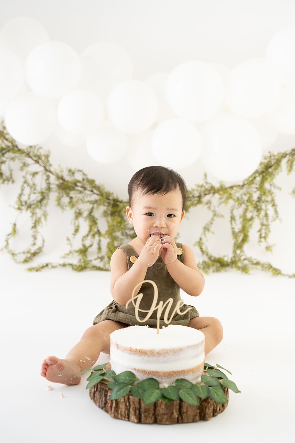 Baby wearing brown "One" outfit sits with a simple white cake on rustic wood slice with greenery garland backdrop.