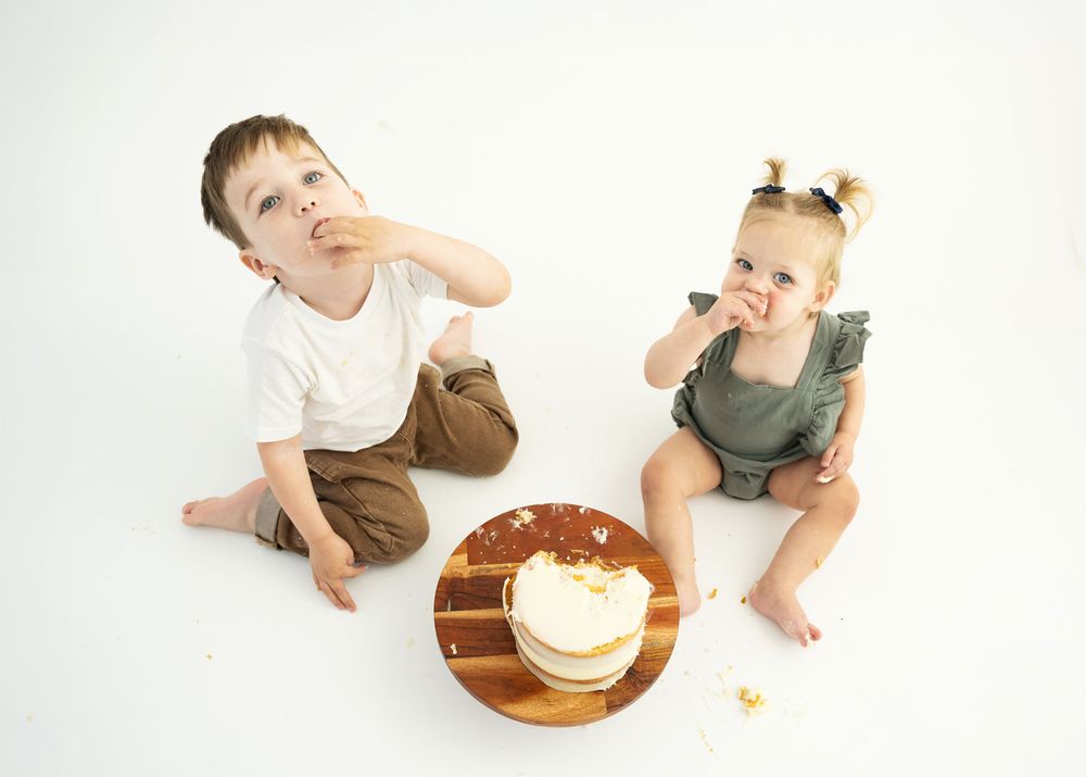 Two children sit on a white studio floor smashing a cake on a wooden cake stand.