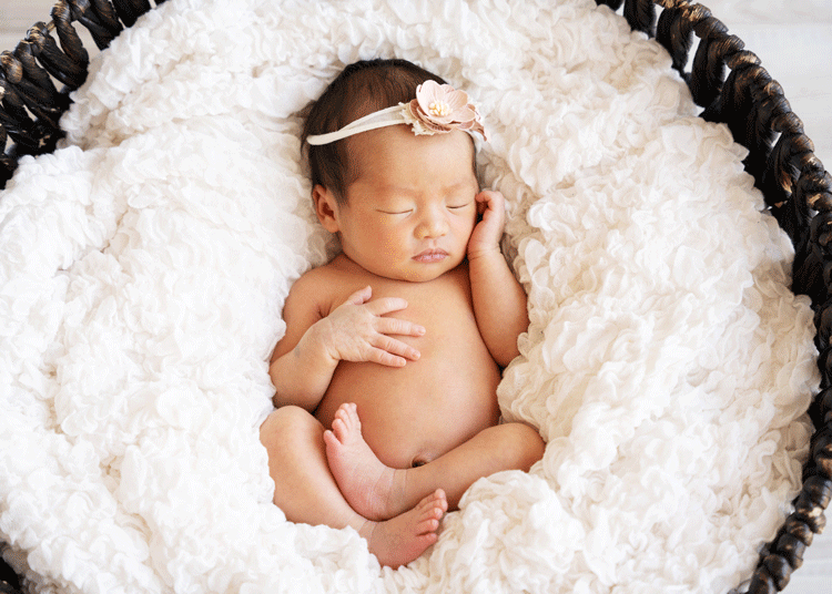 Sleeping newborn baby lying on soft white blanket wearing delicate headband in professional studio portrait.