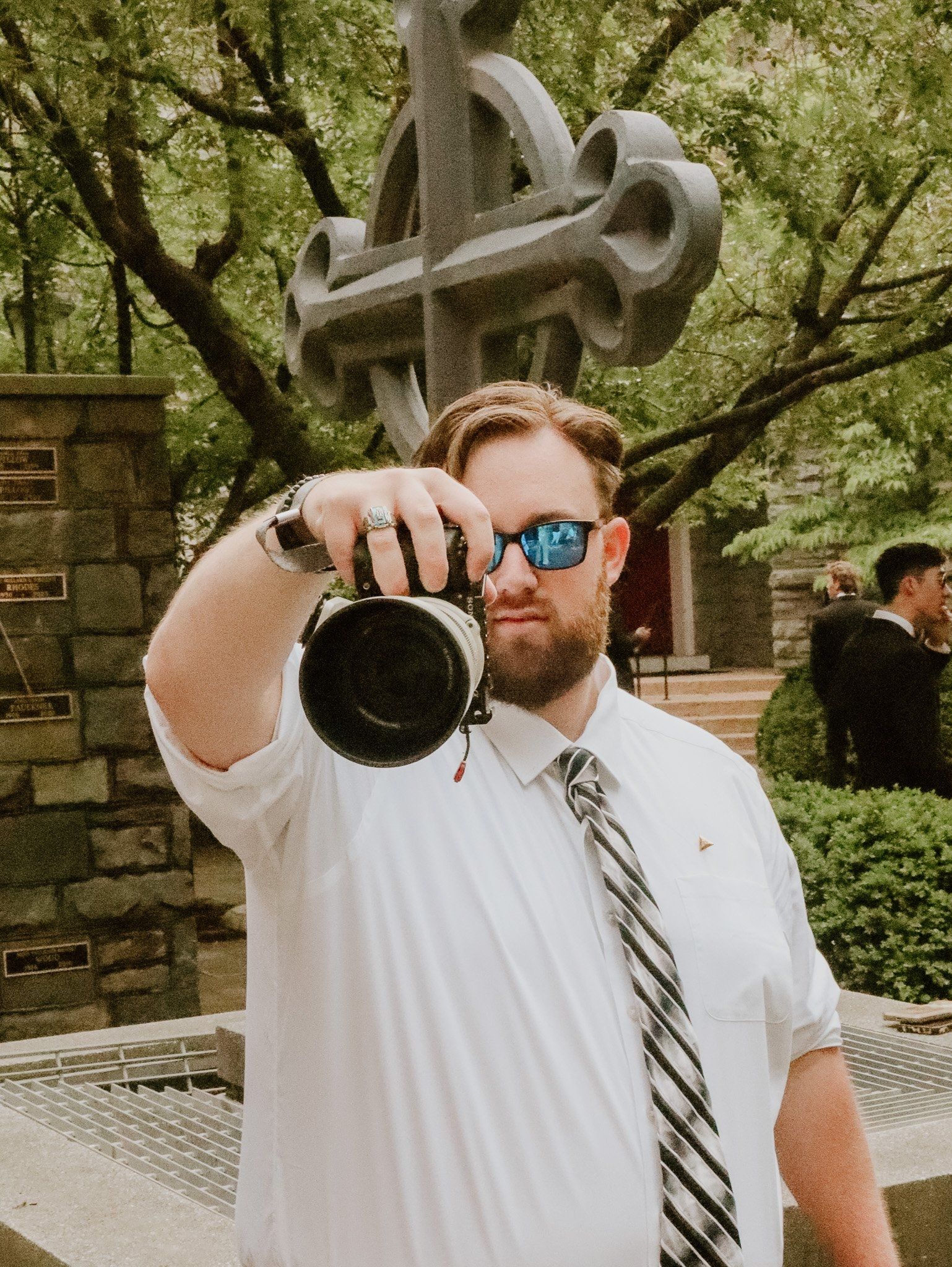 Photographer in white shirt and tie takes photo in front of stone cross monument.