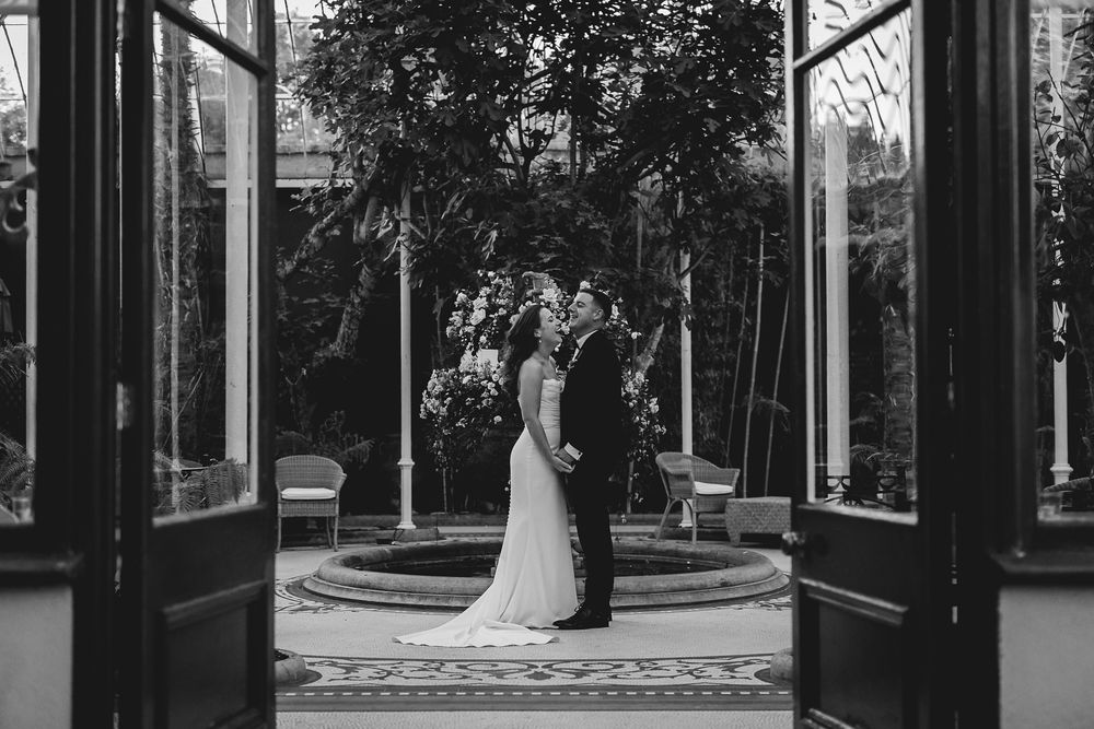 Black and white photo of couple framed by glass doorway in garden setting.