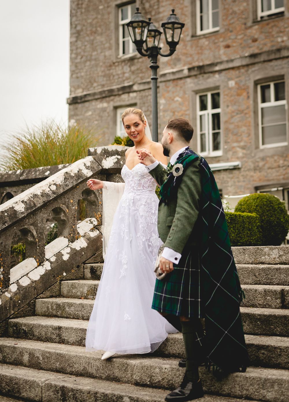 A wedding couple poses on stone steps outside a mansion, with the groom wearing a traditional Scottish kilt.