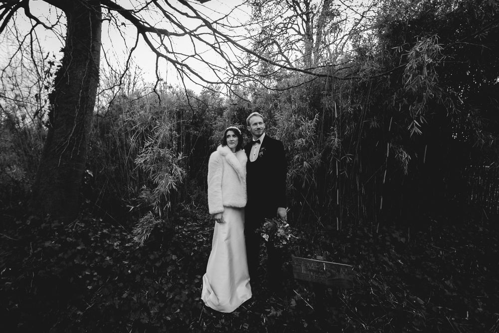 Black and white portrait of newlyweds standing among trees in a moody forest setting.