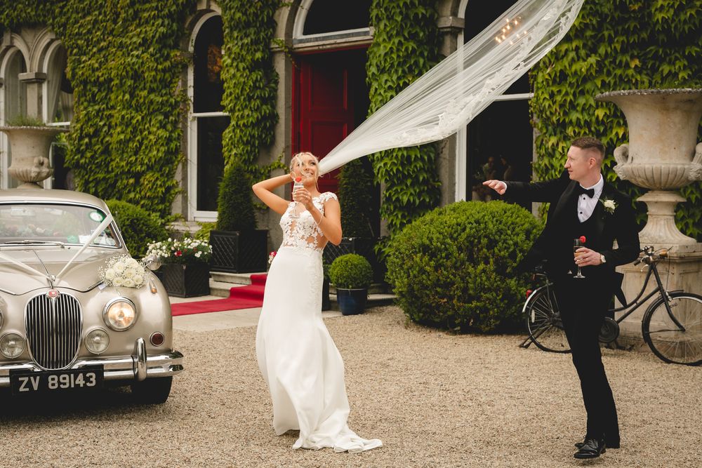 White veil billows dramatically in the wind during wedding photoshoot next to classic car outside ivy-covered venue.