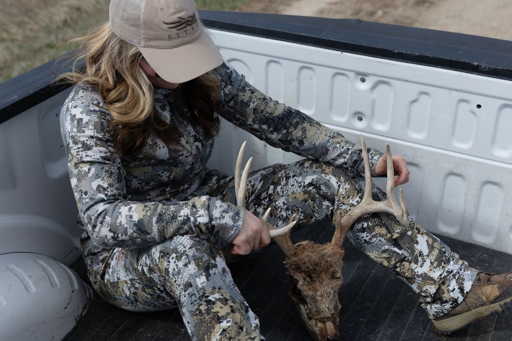 blonde haired girl wearing hat and camo clothing sitting in bed of truck holding whitetail deer skull with antlers