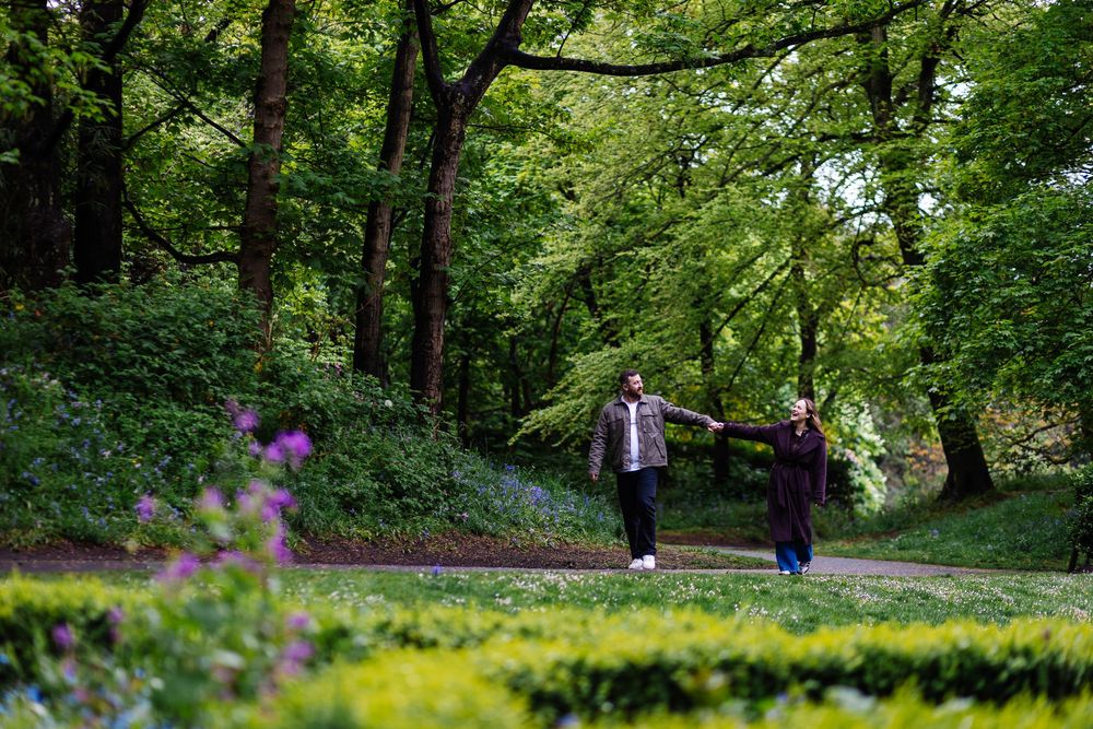 A couple walk hand-in-hand along a park path surrounded by tall trees and blooming flowers at Hesketh Park, Southport by Lynette Matthews