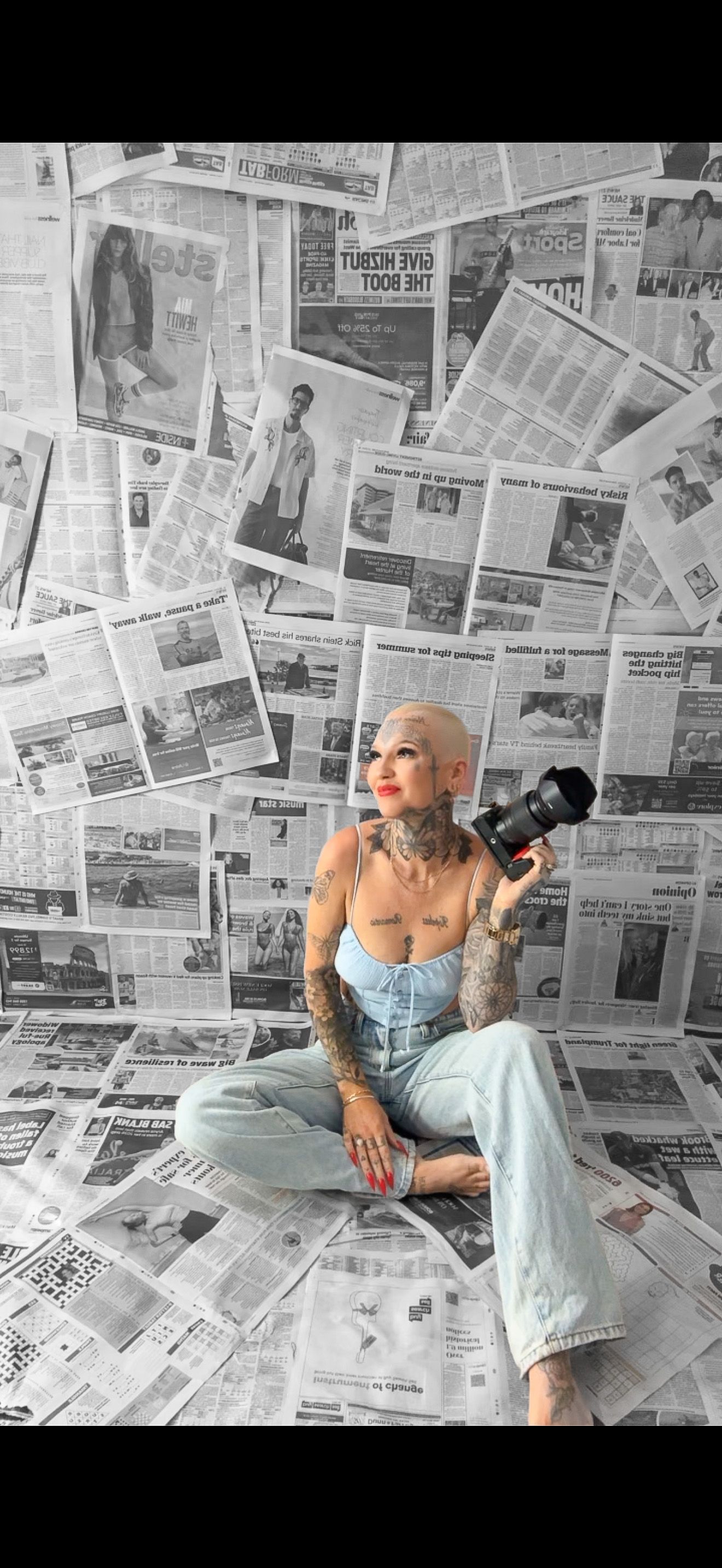 Photographer with tattoos sits cross-legged against wall of newspapers holding camera in casual denim outfit.