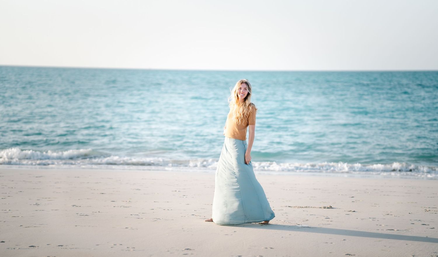 A flowing white dress moves in the ocean breeze on a sandy beach at sunset.