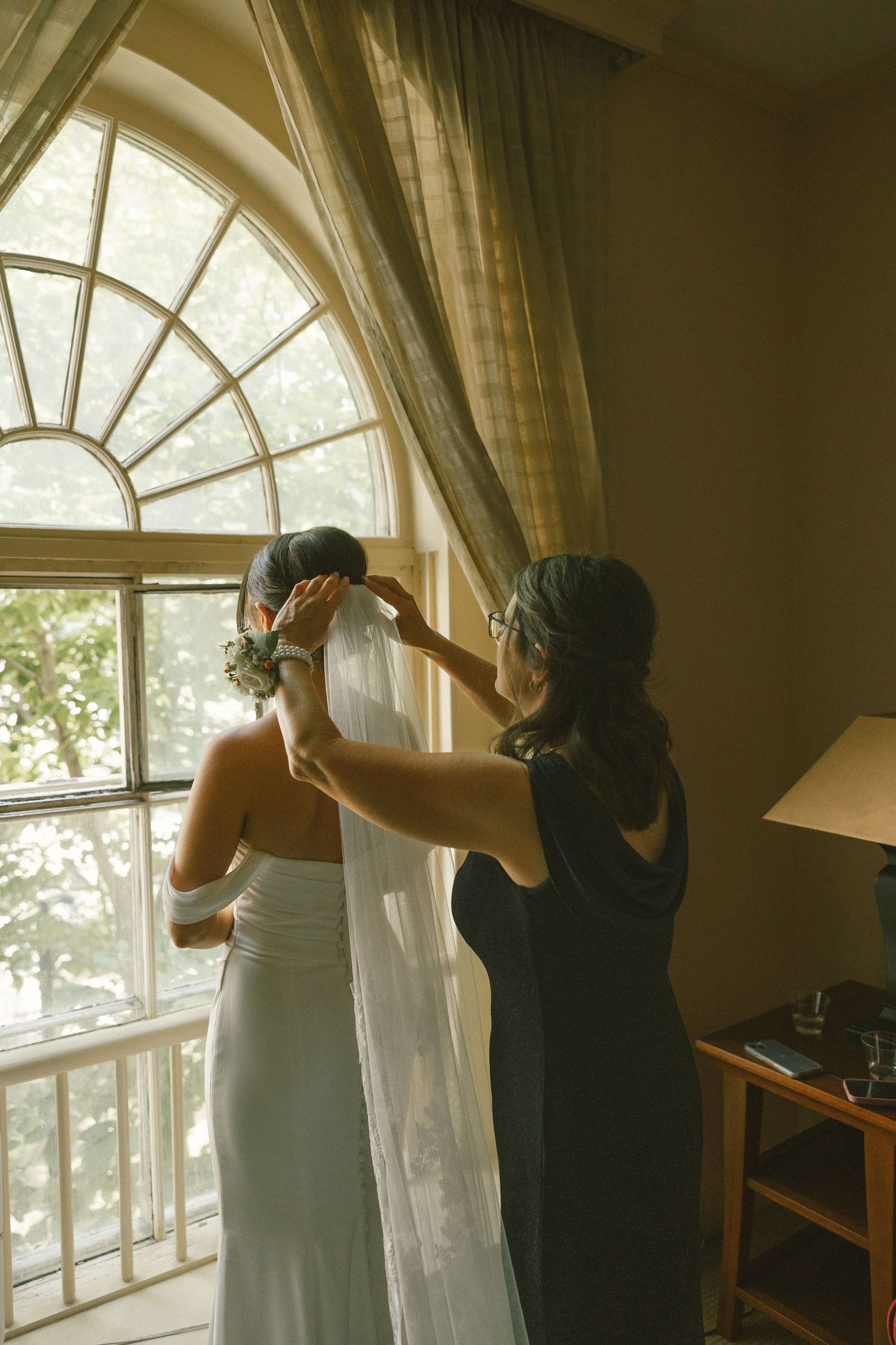 Two people adjust white curtains in front of large arched windows in a sunlit room.